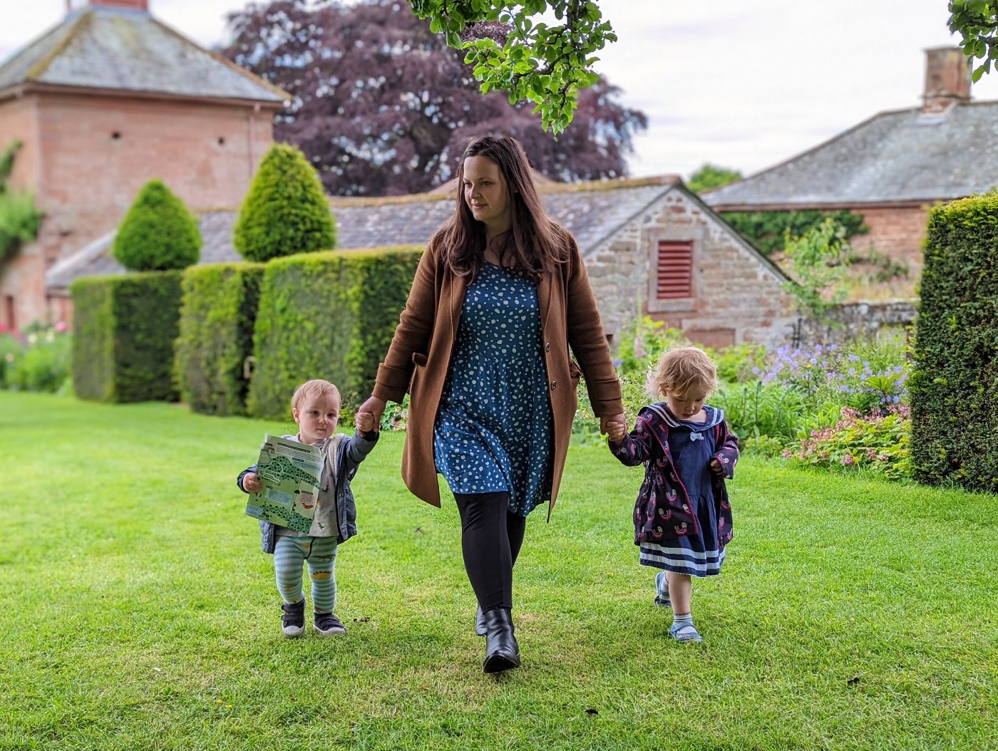 Julia walking hand in hand with two small children across the lawn of the walled garden at Acorn Bank, Temple Sowerby, with clipped hedges and red sandstone buildings behind.