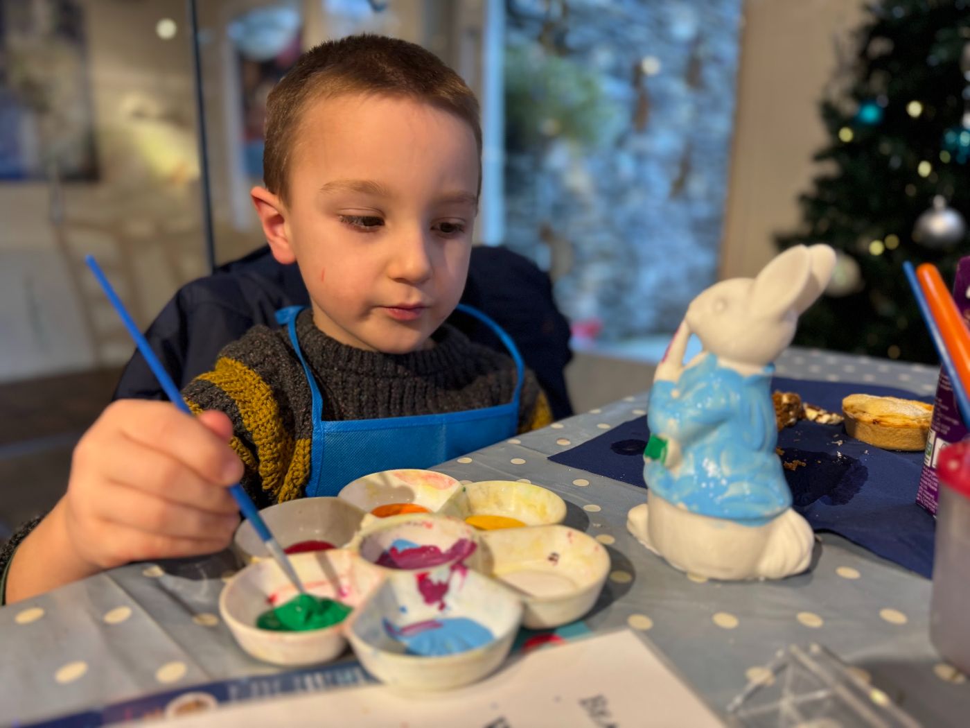 A boy in an apron painting a small white Peter Rabbit figure blue, with a palette of paints and a Christmas tree behind.