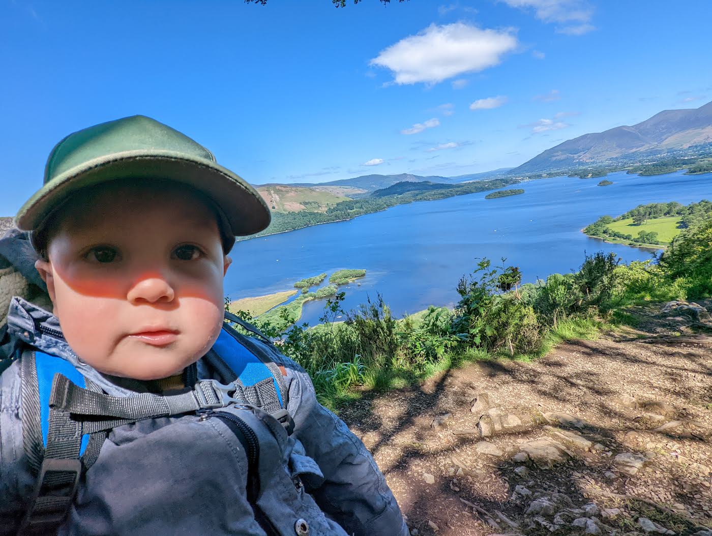 A toddler in a green sun hat in a back carrier, with Derwentwater, its islands and the fells spread out far below.